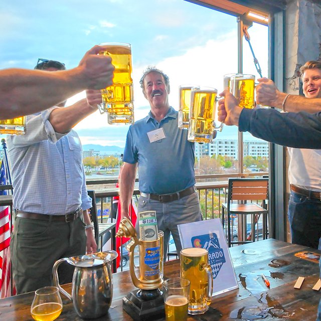 Competitors raising beer steins at the ICSC ColoradoFest Stein Hoisting Competition, with the stein trophy on the table
